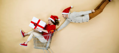 Girl In Shopping Cart With A Package Wrapped In White And A Red Ribbon And A Boy With A Had Flying Behind With Hands On Card Handle