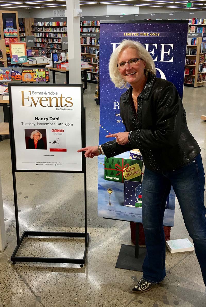 A photograph of Nancy M. Dahl pointing to the sign for her book launch at Barnes and Noble.